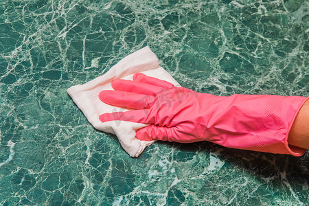 家庭主妇的手戴着粉红色的家用手套，在厨房里用白布擦拭绿色大理石表面