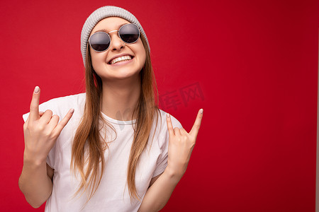 摇滚手势摄影照片_年轻情绪积极、快乐、微笑、迷人的深色金发女性的特写镜头，她的情感真诚，穿着休闲白色 T 恤、灰色帽子和太阳镜，红色背景与复制空间隔离，并展示摇滚手势