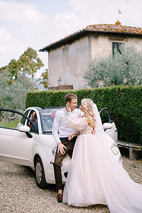 红色敞篷的汽车摄影照片_美丽的新娘和新郎靠在一辆敞篷车上，在意大利佛罗伦萨附近托斯卡纳的老别墅里互相看着对方