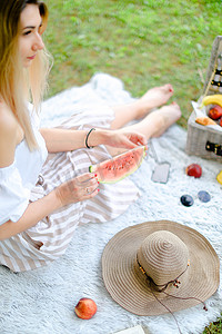 年轻的金发女人坐在靠近水果和帽子的格子上，吃着西瓜，背景是草。