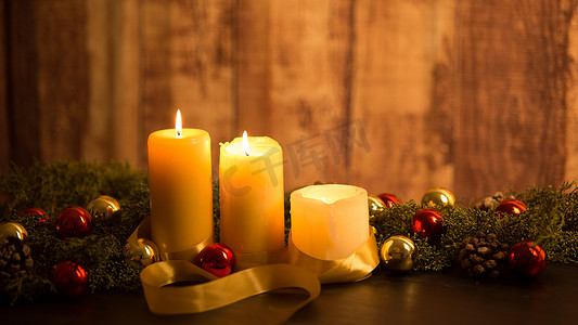 圣诞节概念的温暖：深色木桌上点着三支蜡烛，乡村环境中有松枝、天然松果、金色和红色明亮的小玩意，上面有金色缎带