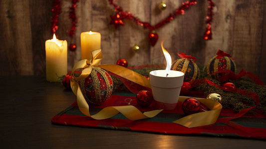圣诞桌布上点着大火焰的蜡烛，周围有松树枝、剪纸小饰物、点燃的蜡烛和悬挂在木制背景上的圣诞装饰，具有散景效果