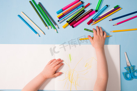 一个小孩用铅笔在相册、复印空间、顶视图、蓝色背景、铅笔、蜡笔、剪刀和孩子的手在桌子上画画