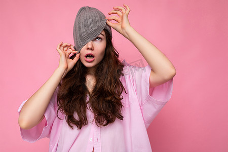 开心心情摄影照片_照片中迷人的疯狂情绪化的黑发年轻女性穿着粉色衬衫和灰色帽子，与世隔绝地站在粉色背景墙上，玩得开心