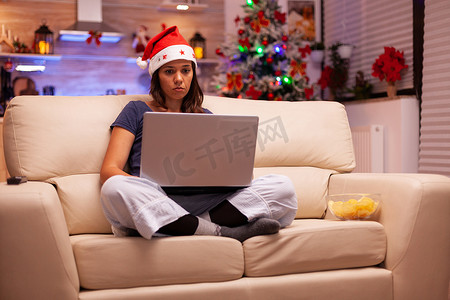 戴着圣诞帽的女人躺在沙发上，用笔记本电脑阅读商务电子邮件