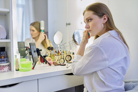 化妆台摄影照片_年轻女子坐在化妆台前，拿着镜子化妆和发型