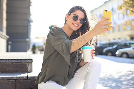 微笑的美丽黑发女人在街上散步，用手机自拍，喝咖啡。