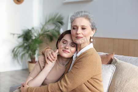 性格开朗的年轻女子坐在家里的沙发上，闭着眼睛拥抱着她的中年母亲，抚摸着脸颊。