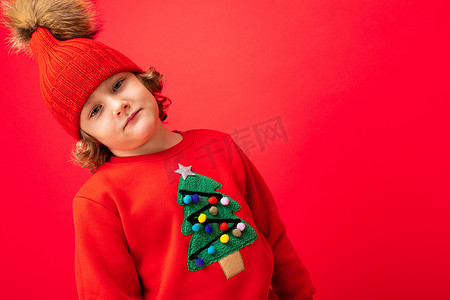 戴着温暖帽子和毛衣的金发酷小孩，红色背景上挂着圣诞树，圣诞概念