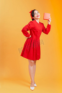人们、圣诞节、生日和假期的概念-穿着红色连衣裙的快乐年轻女人在橙色背景下展示/拿着礼品盒。