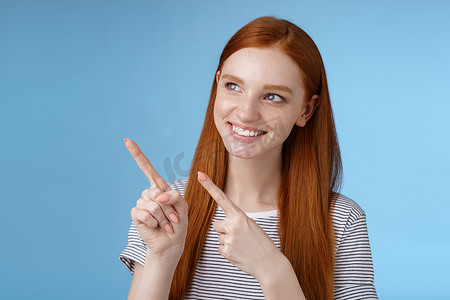 产品宣传摄影照片_迷人的红发女模特纯洁的皮肤姜长发指向左看感兴趣微笑满意表示有趣的广告对使用新产品感兴趣，蓝色背景