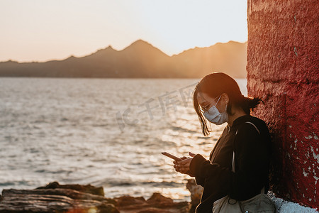 年轻女子在西班牙海岸用他的手机，以巨大的日落为背景，同时使用面具放松概念