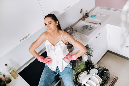 打扫完房子后，一个戴着粉红色手套的家庭主妇女孩疲倦地坐在厨房里。在白色的厨房里，这个女孩洗了盘子，正在休息。很多洗过的盘子