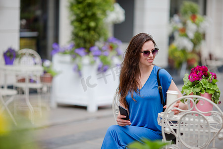 快乐的年轻都市女性，在欧洲的户外咖啡馆里拥有现代手机和咖啡。