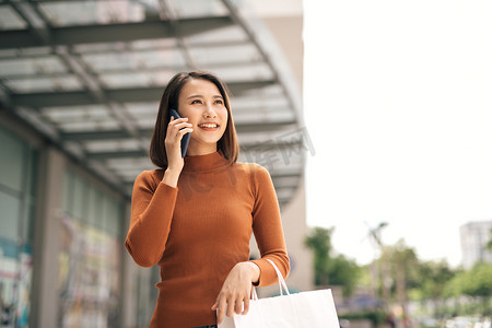 商场购物袋摄影照片_快乐的亚洲年轻女性在手机上聊天，提着纸购物袋在户外商场散步的画像