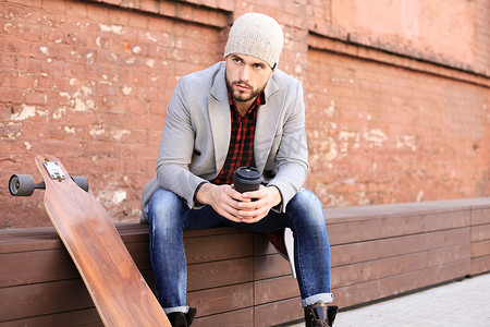 英俊的年轻人穿着灰色外套和帽子，休息，坐在长板上。