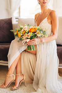 凉鞋摄影照片_一位穿着婚纱和高跟凉鞋的新娘坐在房间的沙发上，手里拿着一束婚礼花束