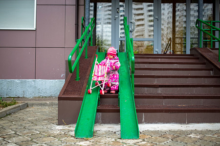 可爱设施摄影照片_可爱的幼儿带着玩具婴儿车沿着轮椅、手推车和婴儿车的钢栏杆坡道行走。