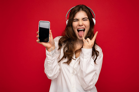 身穿红色背景墙上突显的白衬衫的迷人正能量年轻黑发卷发女性拿着并展示手机，手机上的显示屏空空如也，戴着白色蓝牙耳机听音乐，展示摇滚手势和舌头