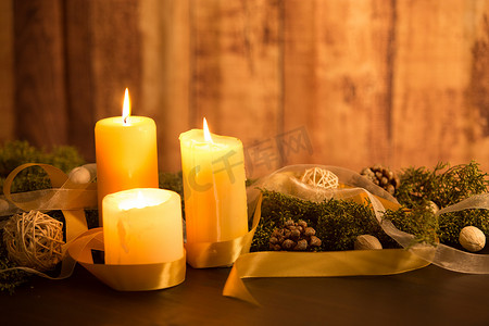 圣诞节概念的温暖：深色木桌上点燃了三支蜡烛，乡村风格的木制环境配有松枝、天然松果、金色缎子和白色欧根纱丝带