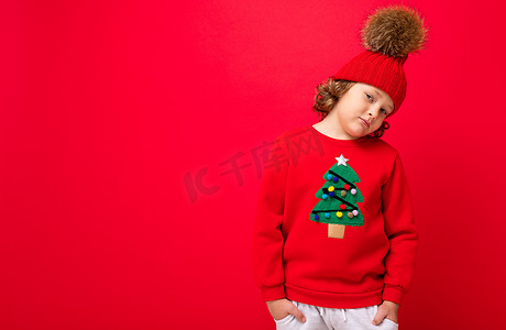 戴着温暖帽子和毛衣的金发酷小孩，红色背景上挂着圣诞树，圣诞概念