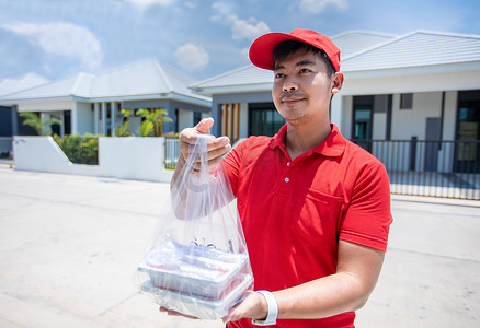 买送摄影照片_亚洲送货员穿着红色制服，戴着红色帽子，拿着塑料袋里的食物盒，将食物送到家门口的顾客手中。
