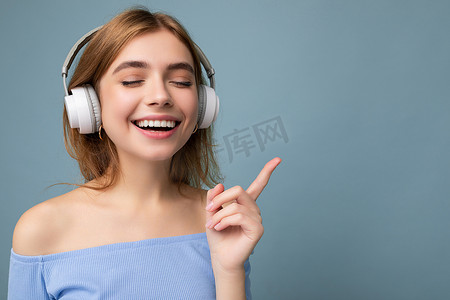 特写照片：美丽正面微笑的年轻金发女子身穿蓝色裁剪上衣，与蓝色背景墙隔开，戴着白色无线蓝牙耳机听酷音乐并享受