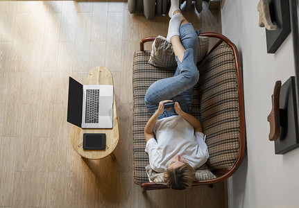 一位女士躺在沙发上使用智能手机，桌上放着笔记本电脑和电子阅读器。