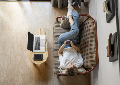 一位女士躺在沙发上使用智能手机，桌上放着笔记本电脑和电子阅读器。