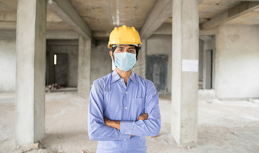 covid-19大流行后重返工作岗位、开放建筑工地的概念 — 自信的建筑工人戴着建筑头盔，双臂交叉在现场的医用口罩。