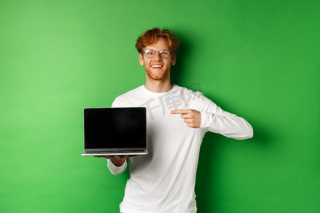 戴着眼镜和白色长袖 T 恤的快乐红头发人，手指指着空白的笔记本电脑屏幕，微笑着，站在绿色背景上