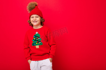 小孩圣诞老人摄影照片_戴着温暖帽子和毛衣的金发酷小孩，红色背景上挂着圣诞树，圣诞概念