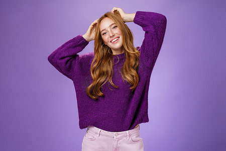 紫色背景炫酷摄影照片_穿着紫色保暖毛衣的无忧无虑、快乐而酷炫的时尚红发女学生享受假期的伸展、双手靠近头部、倾斜和微笑，在紫色背景下愉快地放松