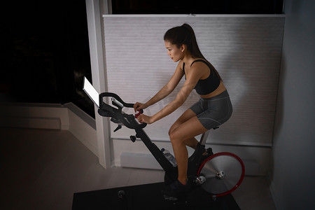 家庭锻炼室内固定自行车亚洲女孩自行车屏幕与在线课程女性在室内智能健身设备上进行自行车运动训练。