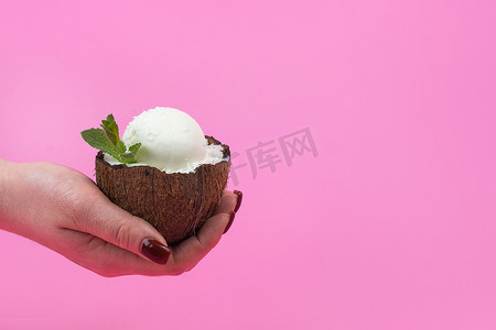 白色冰淇淋球摄影照片_香草冰淇淋球放在新鲜椰子的一半里，手上装饰着粉红色背景的薄荷叶