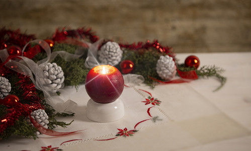 圣诞节设置：前景上有交叉屏幕效果的红色点燃蜡烛，周围环绕着松树枝、红色小饰品、红色和白色丝带、圣诞桌布上的白色松果，具有散景效果