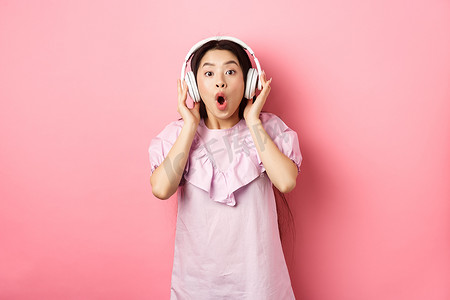 兴奋的亚洲女孩听到美妙的歌曲，戴着耳机听音乐，站在粉红色背景中看起来很惊讶