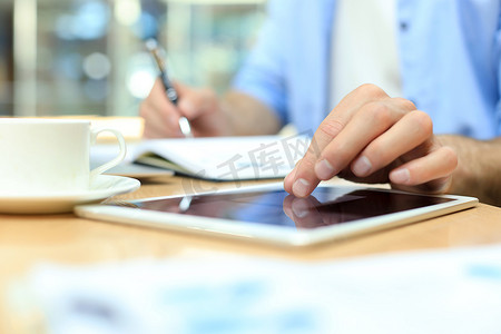 男人用笔在纸质笔记本上写工作计划，在办公桌上的数字平板电脑上浏览互联网。
