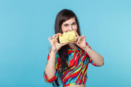 微笑的年轻女子手里拿着一个黄色的老式相机模型，在蓝色背景上摆出姿势，并有复制空间。