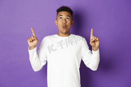 紫色背景炫酷摄影照片_身穿白色运动衫的非洲裔美国人印象深刻，他用惊讶的表情指指点点，展示酷炫的广告，站在紫色背景上