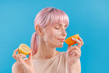 粉红色头发的轻松女人的肖像，手里拿着一半新鲜的橙子和柠檬，闭着眼睛闻着它的味道，与蓝色背景隔离