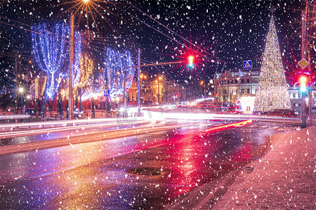 夜城里有圣诞装饰、云杉和移动汽车的车头灯痕迹，倒映在降雪中湿漉漉的道路上。