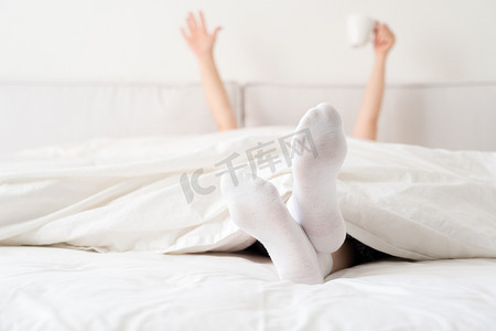 女手从床上的毯子下拿着一杯咖啡。