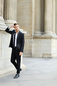 年轻英俊的男子穿着黑色西装和领带靠在建筑柱上。