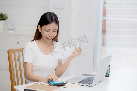 年轻的亚洲女性用家里桌上的计算器计算财务家庭，女孩检查账单并成功节省家庭开支、债务结清、税收和会计、商业理念。