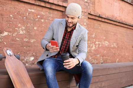 穿着灰色外套和帽子的英俊年轻人使用智能手机，休息，坐在长板上。