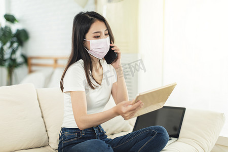 在家工作的年轻女性，戴着口罩保护自己，坐在沙发上使用笔记本电脑进行远程办公，一边看平板电脑一边打电话
