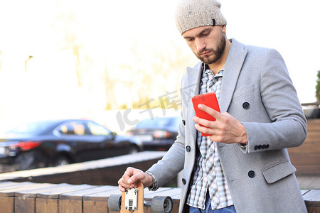 穿着灰色外套和帽子的英俊年轻人使用智能手机休息，站在长板上。
