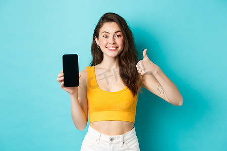 时尚现代女性的肖像高兴地微笑，推荐应用程序，在空的智能手机屏幕上向您展示徽标横幅，竖起大拇指表示批准，赞扬应用程序，蓝色背景