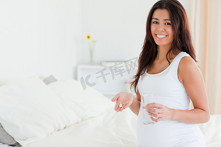 坐在床上拿着一杯水和药丸的漂亮孕妇